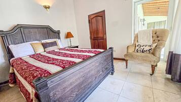 A bedroom with a large bed, a wooden door, a chair with a cushion, and a wall-mounted light fixture.