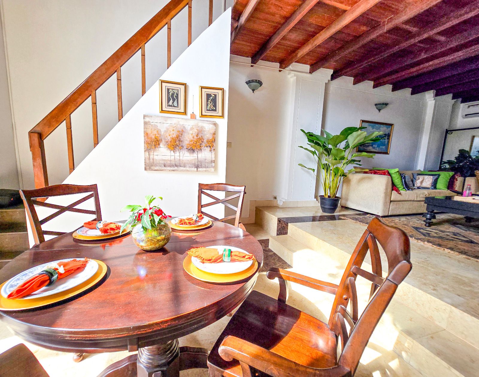 A dining area with a wooden table, chairs, and a staircase leading to a living room with a couch and a plant.