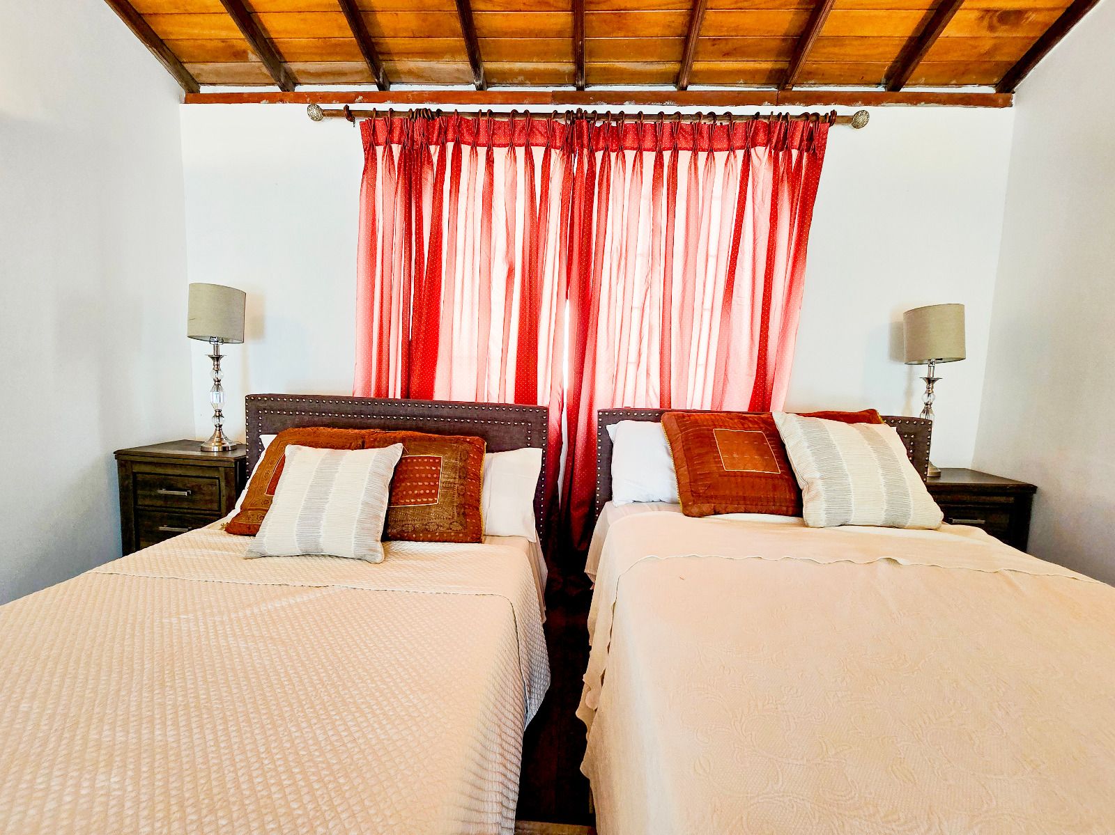 A double bed room with a wooden ceiling, two bedside tables with lamps, and a red curtain.
