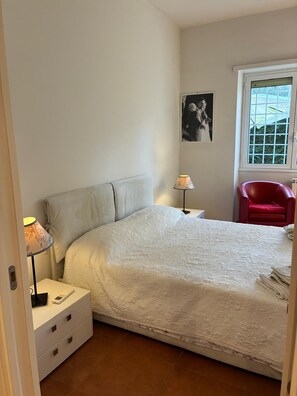 Ein Schlafzimmer mit Bett, Nachttisch, Lampe, Stuhl und einem Fenster mit Blick.