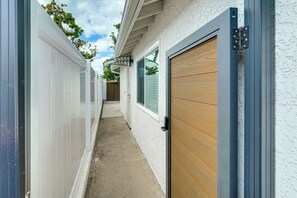A narrow concrete pathway leading to a wooden door with a metal frame, flanked by a white wall and a garage door. - Fullerton Suite w/ Private Yard, Extended Stays (Fullerton)