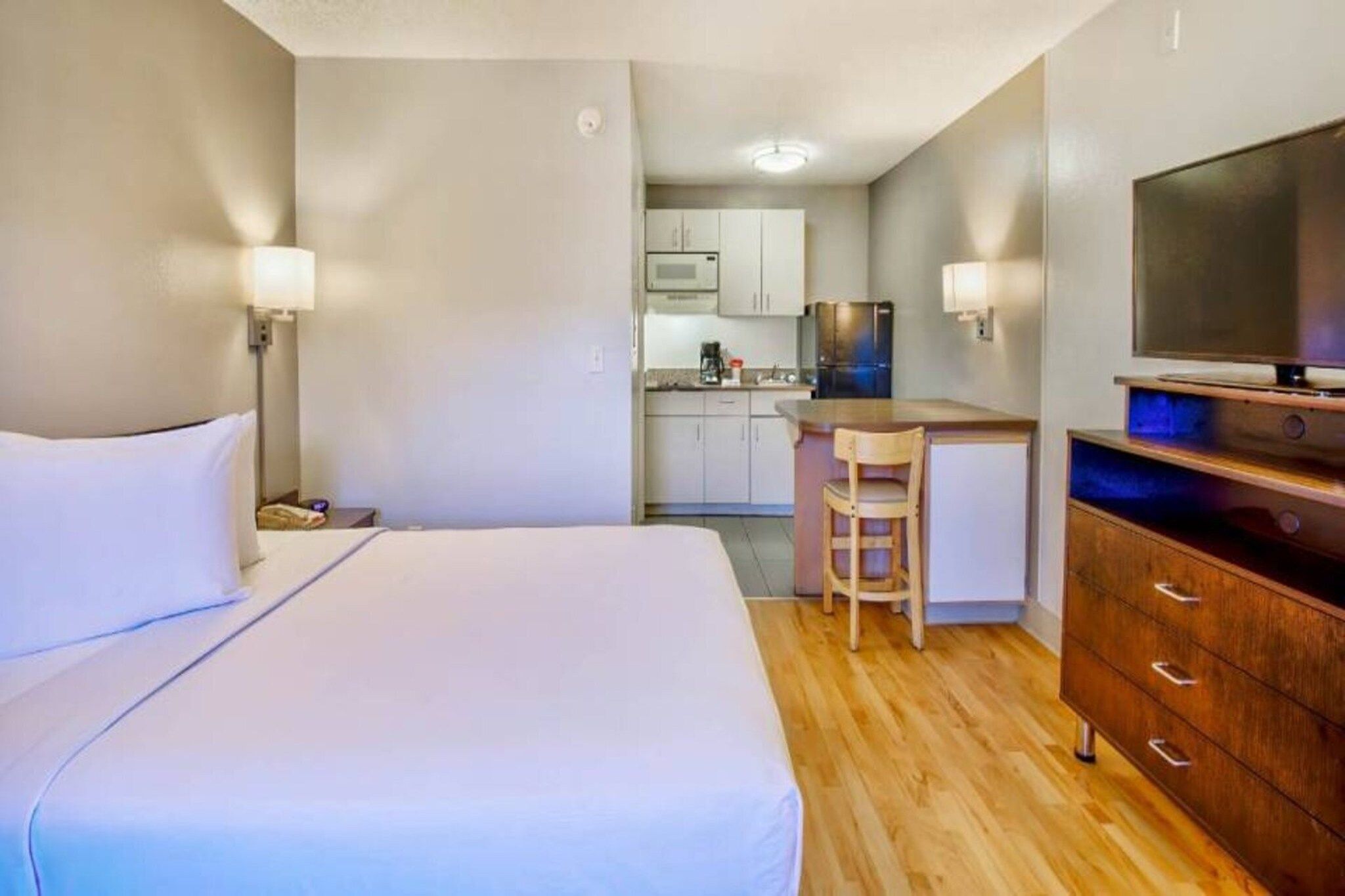 A hotel room with a bed, a desk, a chair, a TV, and a kitchenette in the background.