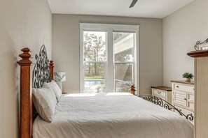 Ein Schlafzimmer mit Bett, Nachttisch, Kommode und Fenster mit Blick auf einen Pool und Bäume.