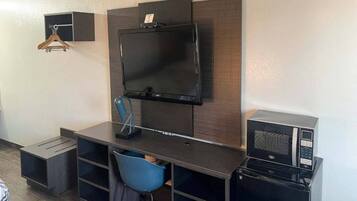 A compact hotel room with a flat-screen TV mounted on a wooden panel, a small desk with a chair, and a microwave on the right.