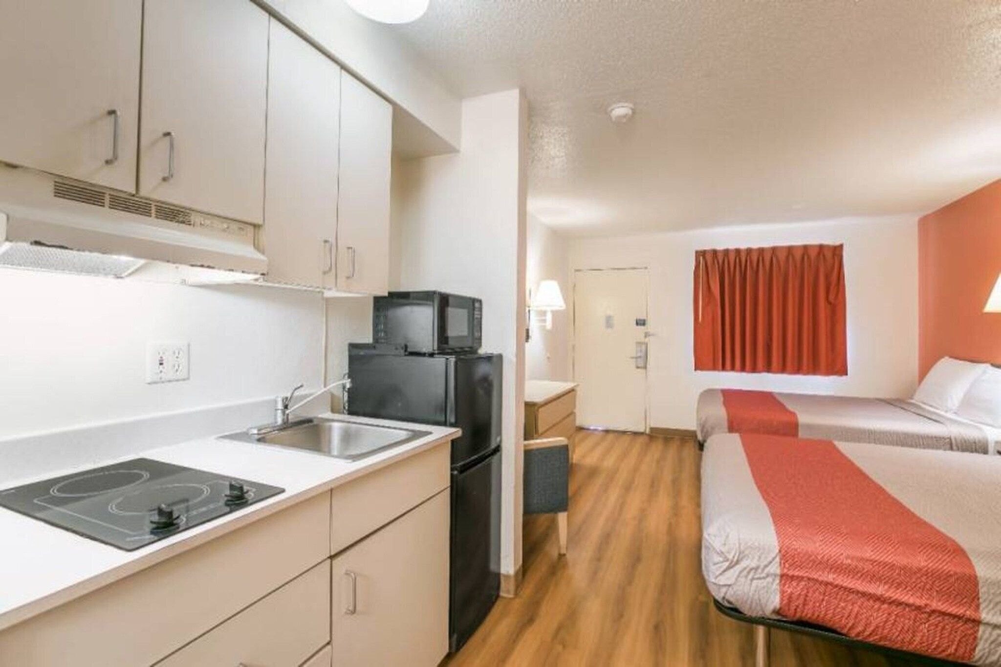Une chambre d’hôtel compacte, dotée d’une kitchenette, d’un lit recouvert d’une courtepointe rouge et blanche, et d’une fenêtre ornée de rideaux rouges.