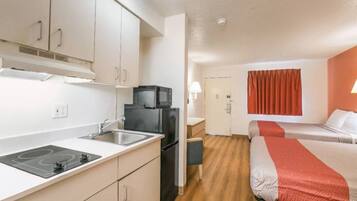 Une chambre d’hôtel compacte, dotée d’une kitchenette, d’un lit recouvert d’une courtepointe rouge et blanche, et d’une fenêtre ornée de rideaux rouges.
