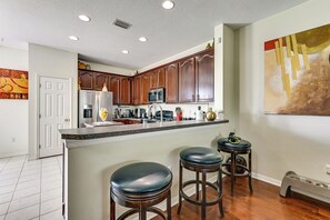 A kitchen with wooden cabinets, a stainless steel refrigerator, a microwave, and a coffee maker. There are two stools and a painting on the wall. - Pool Access! Steps from Upper Tampa Bay Trail (Tampa)