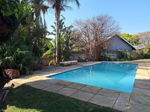 Piscina retangular com borda ladrilhada, rodeada por um pátio de pedra e uma área gramada com diversas plantas e árvores.