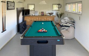Une table de billard avec des billes, un téléviseur et un meuble dans une pièce dotée de fenêtres et d’une plante.