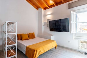 Une chambre à coucher avec un lit, une étagère en métal blanche, un téléviseur fixé au mur et une fenêtre avec des stores.