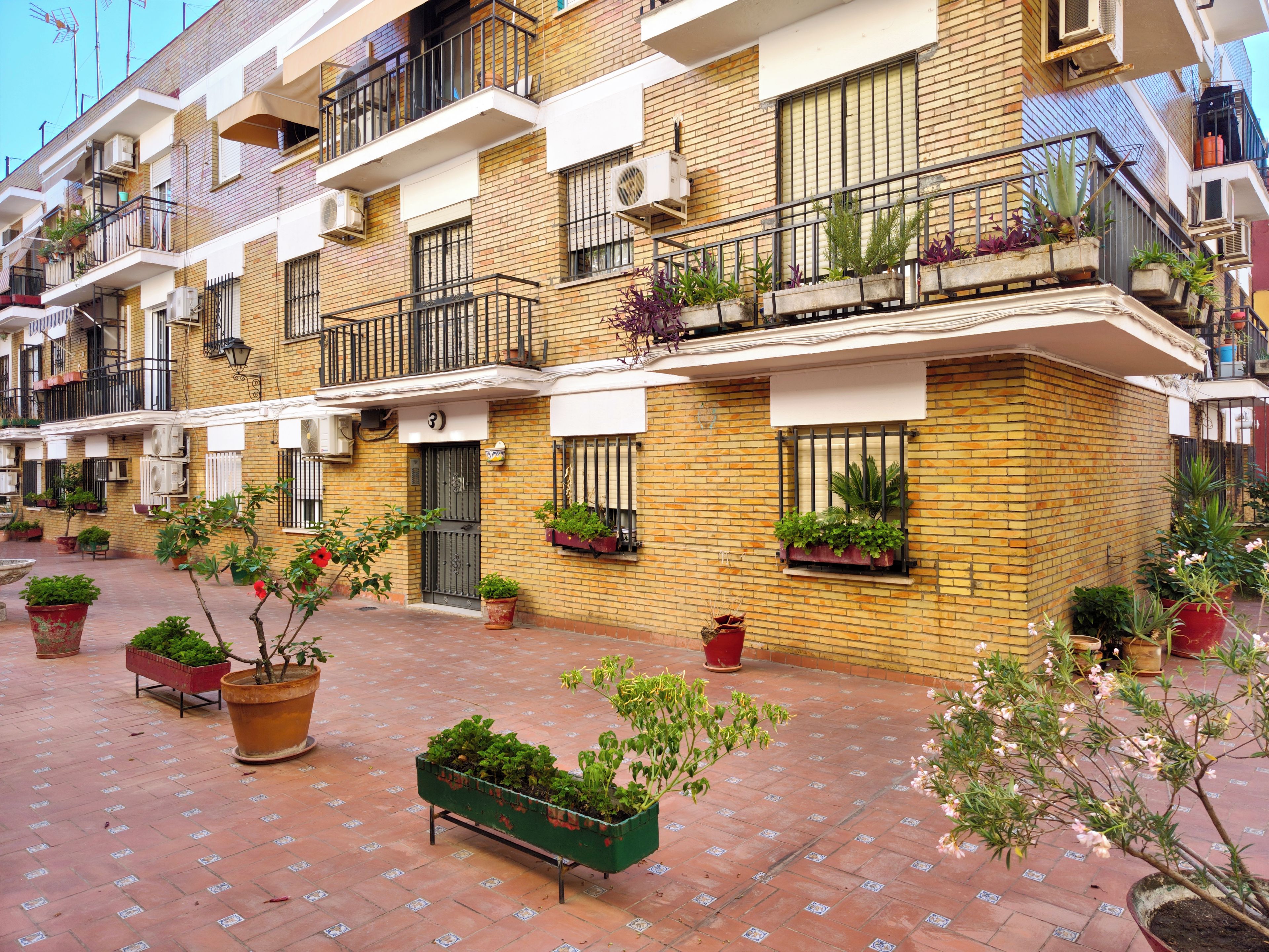 A row of terracotta-brick buildings with balconies, potted plants, and a tiled courtyard.