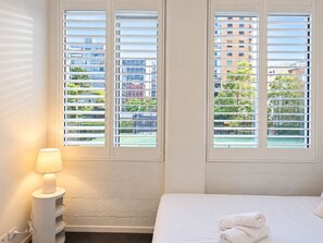 A hotel room with a bed, a nightstand with a lamp, and a view of buildings through the window blinds. - Little Bourke City Charm (Melbourne)