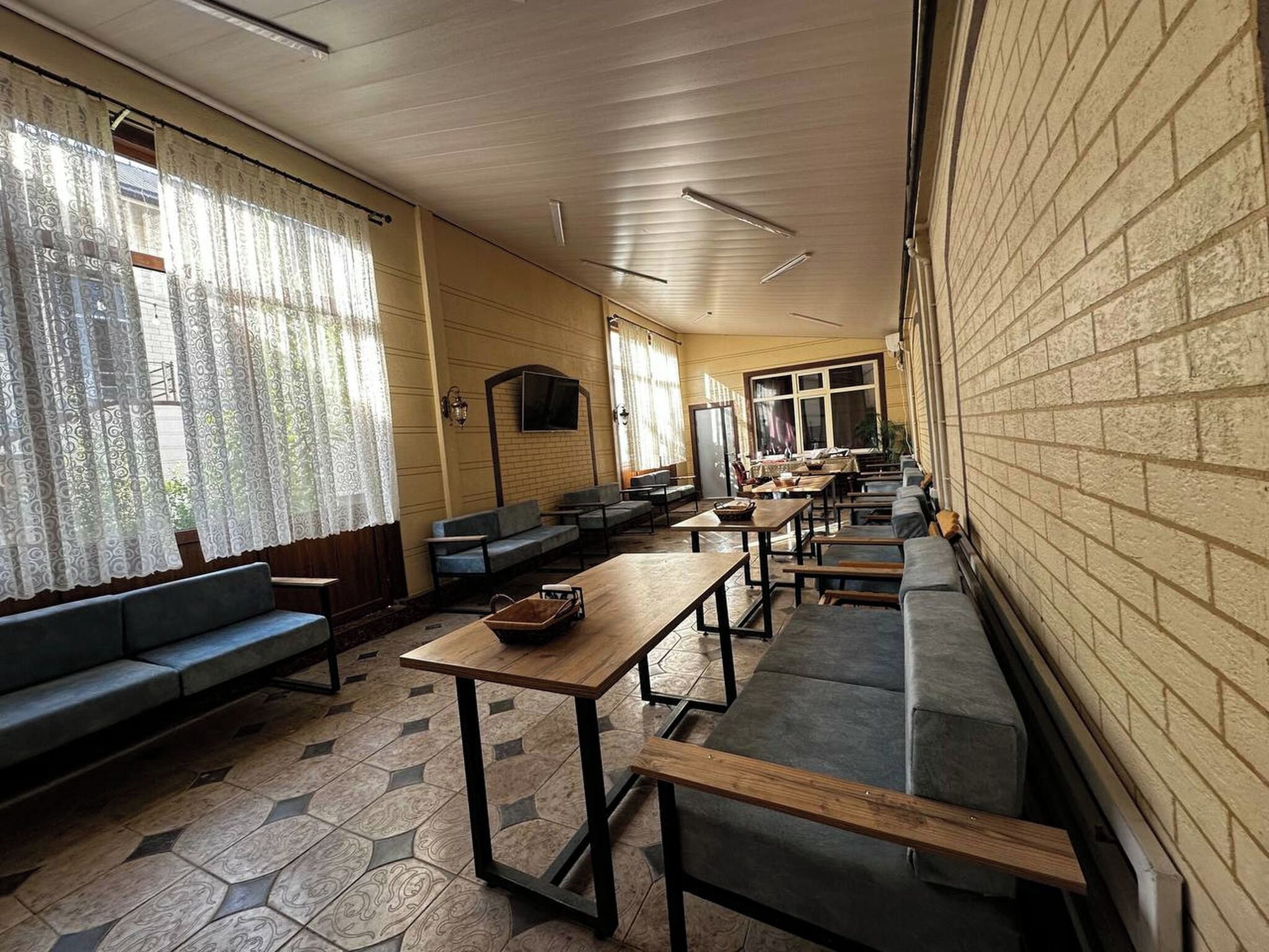 A long corridor with benches and tables, large windows with curtains, and a brick wall on one side.