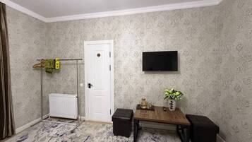 A room with a white door, a mounted TV, a small table with a vase and a remote control, and a radiator.