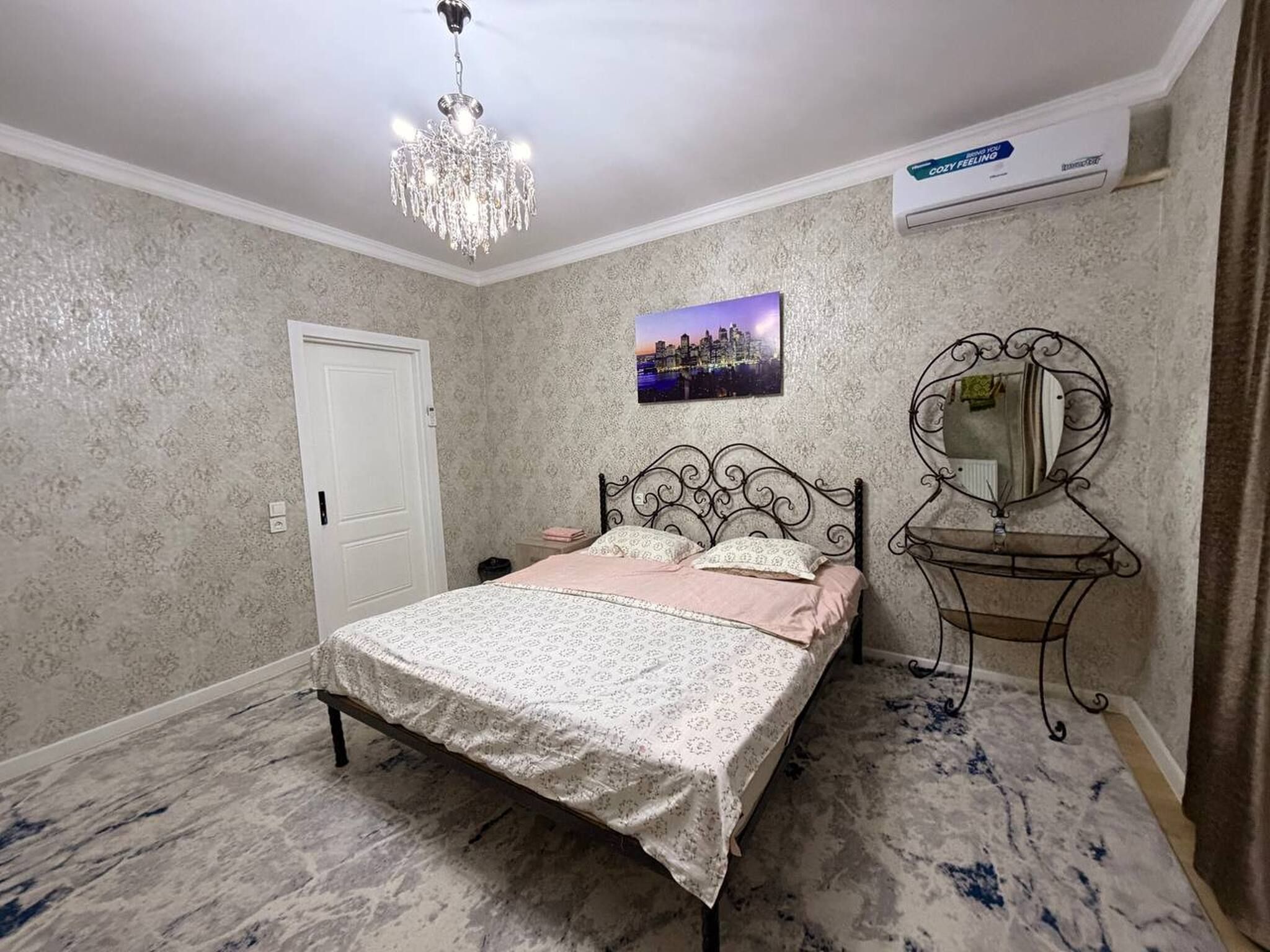 A bedroom with a bed, a chandelier, a cityscape picture, a mirror, and an air conditioning unit.