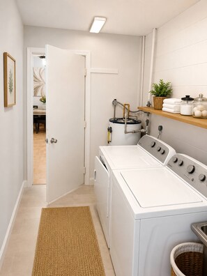 A laundry room with a front-loading washer and dryer, a shelf with towels and a plant, and a door leading to another room. - Stay at Our Beautiful Home in Kissimmee (Kissimmee)
