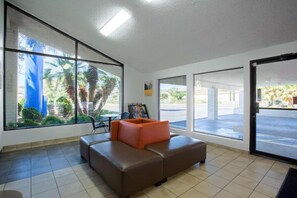 A waiting area with a brown sofa and orange cushions, large windows with a view of palm trees, and a tiled floor. - 2 Full Beds | Micfridge (Kingman)