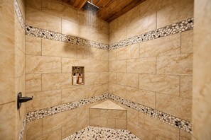 A shower with beige tiles and pebble accents, a showerhead, and a bottle of shampoo. - Broken Bow Lodge: Hot Tub, Sleeps 18 (Broken Bow)