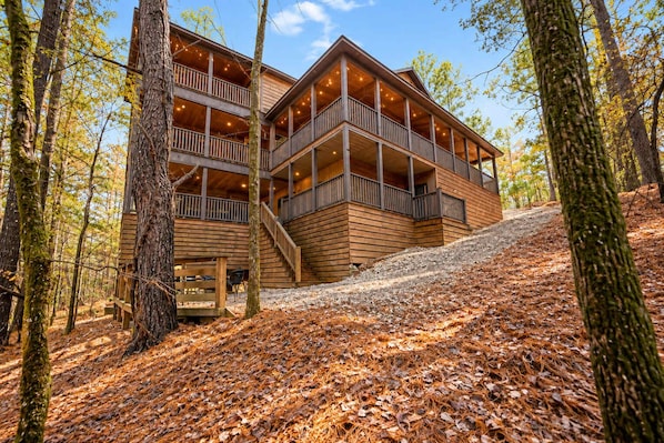 A multi-story wooden cabin with balconies, surrounded by trees and a forest floor covered with fallen leaves. - Broken Bow Lodge: Hot Tub, Sleeps 18 (Broken Bow)