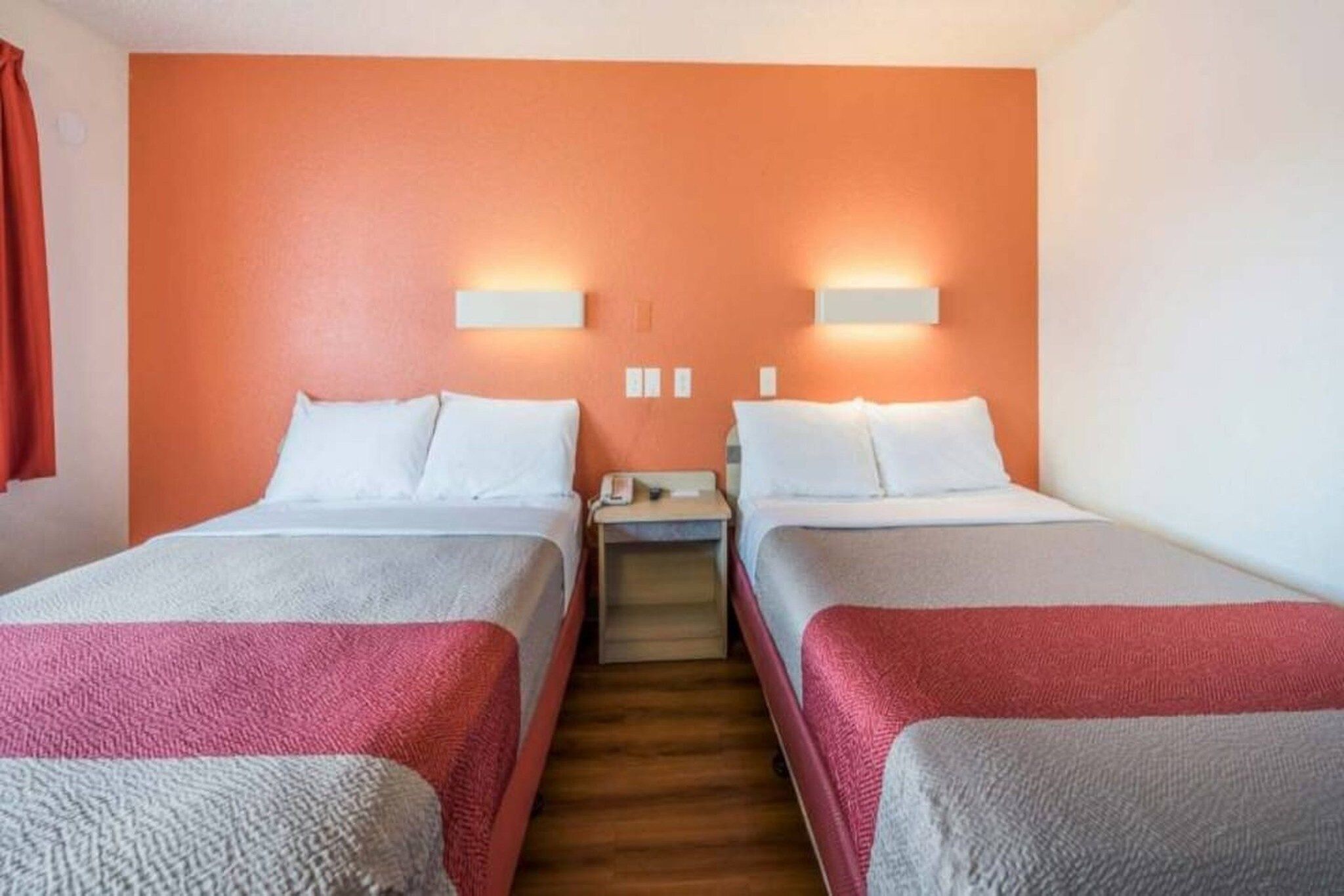 A hotel room with two beds, each with white linens and red blankets. There are two wall-mounted light fixtures on the orange wall.