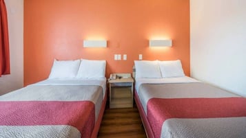 A hotel room with two beds, each with white linens and red blankets. There are two wall-mounted light fixtures on the orange wall.