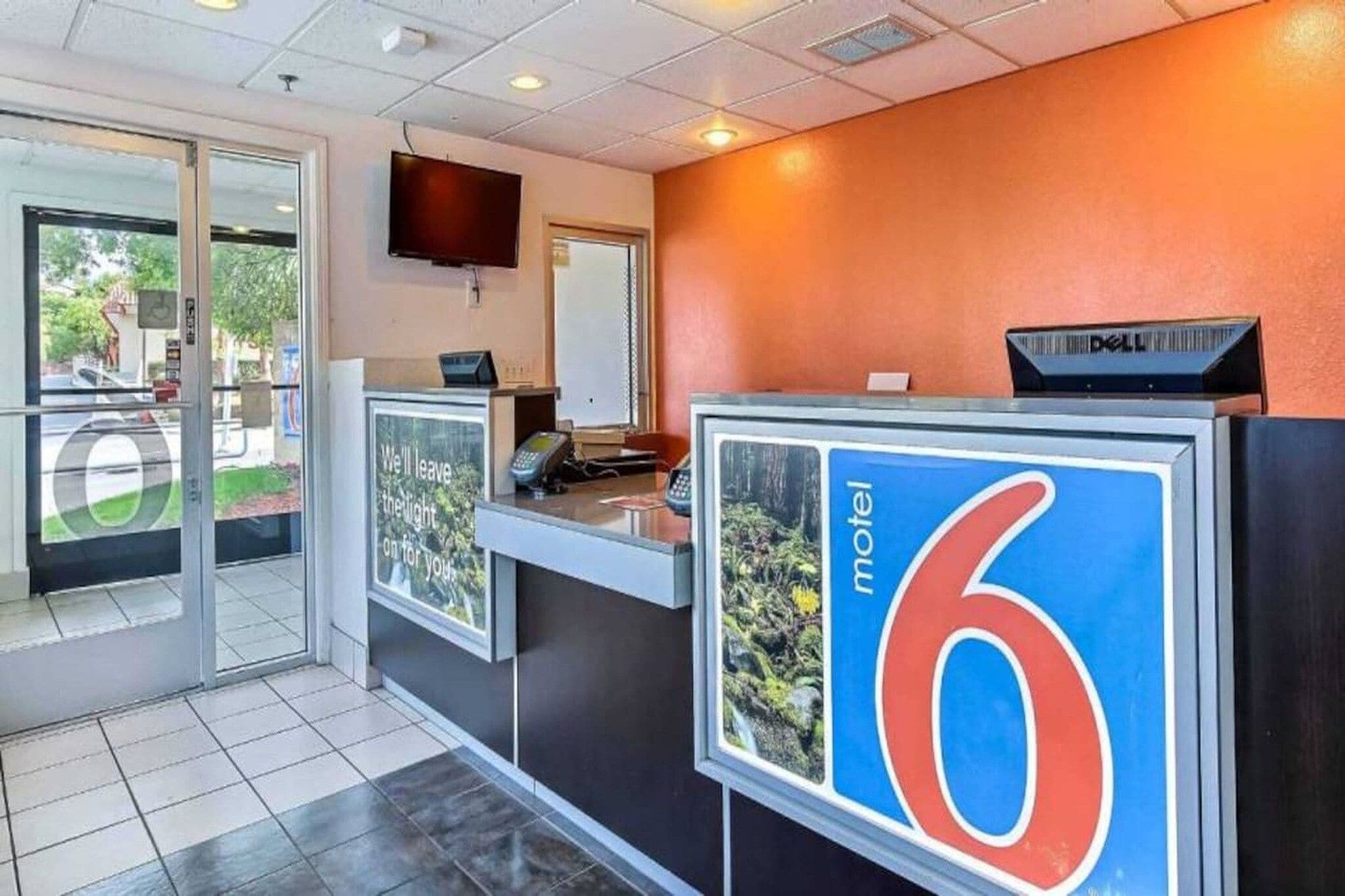 A reception desk with a large sign indicating room number 6, a flat-screen TV mounted on the wall, and a Dell computer on top of the desk.