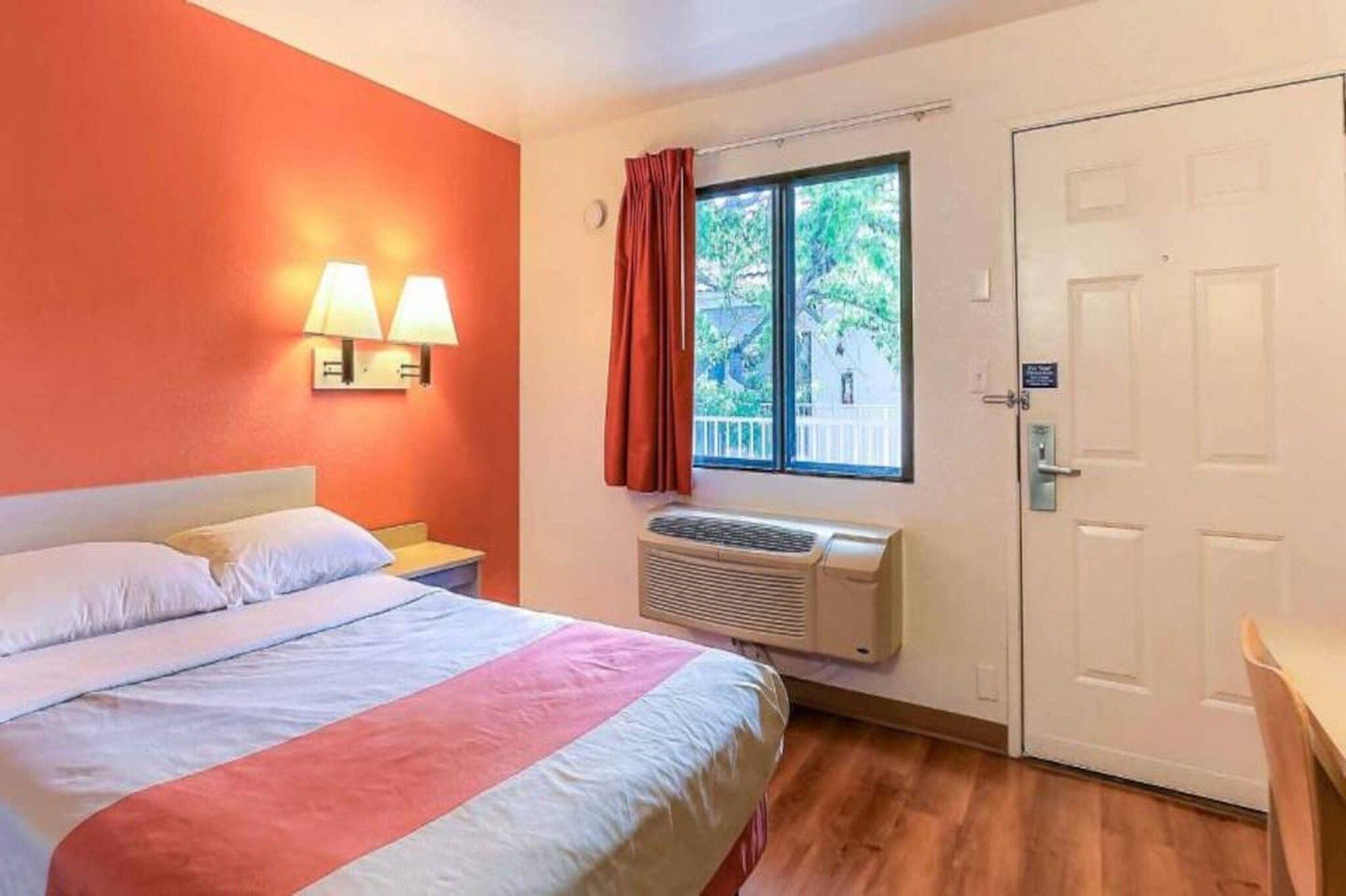 A hotel room with a bed, a nightstand, a window with curtains, an air conditioning unit, and a door.