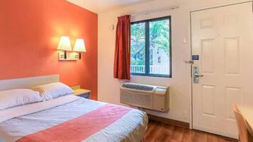 A hotel room with a bed, a nightstand, a window with curtains, an air conditioning unit, and a door.