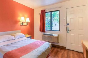 Una habitación de hotel con cama, mesita de noche, ventana con cortinas, unidad de aire acondicionado y puerta.