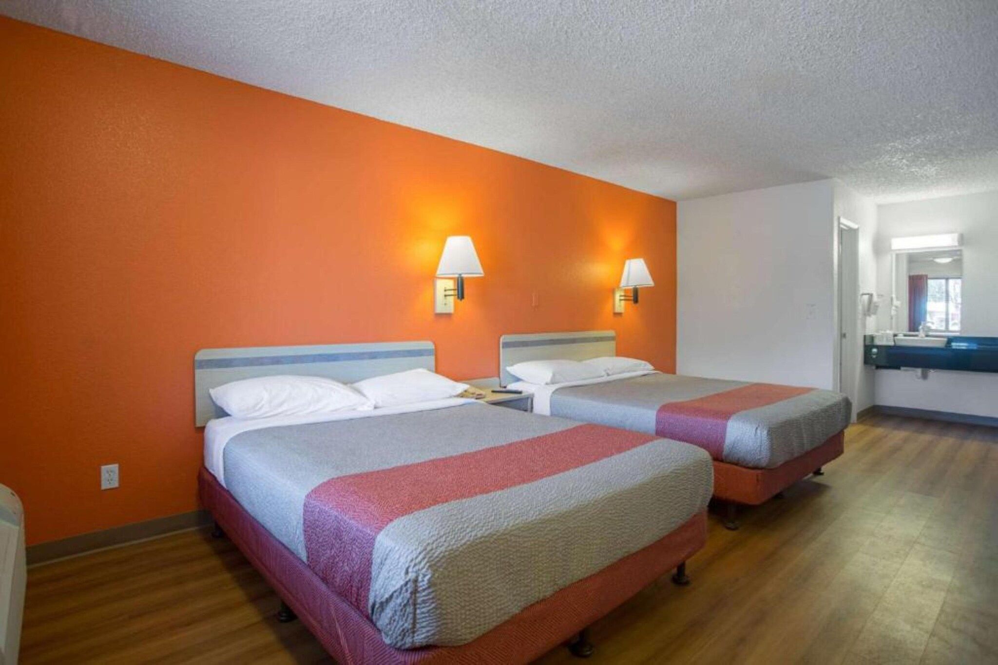 Habitación de hotel con dos camas, paredes anaranjadas y baño con espejo y lavamanos.