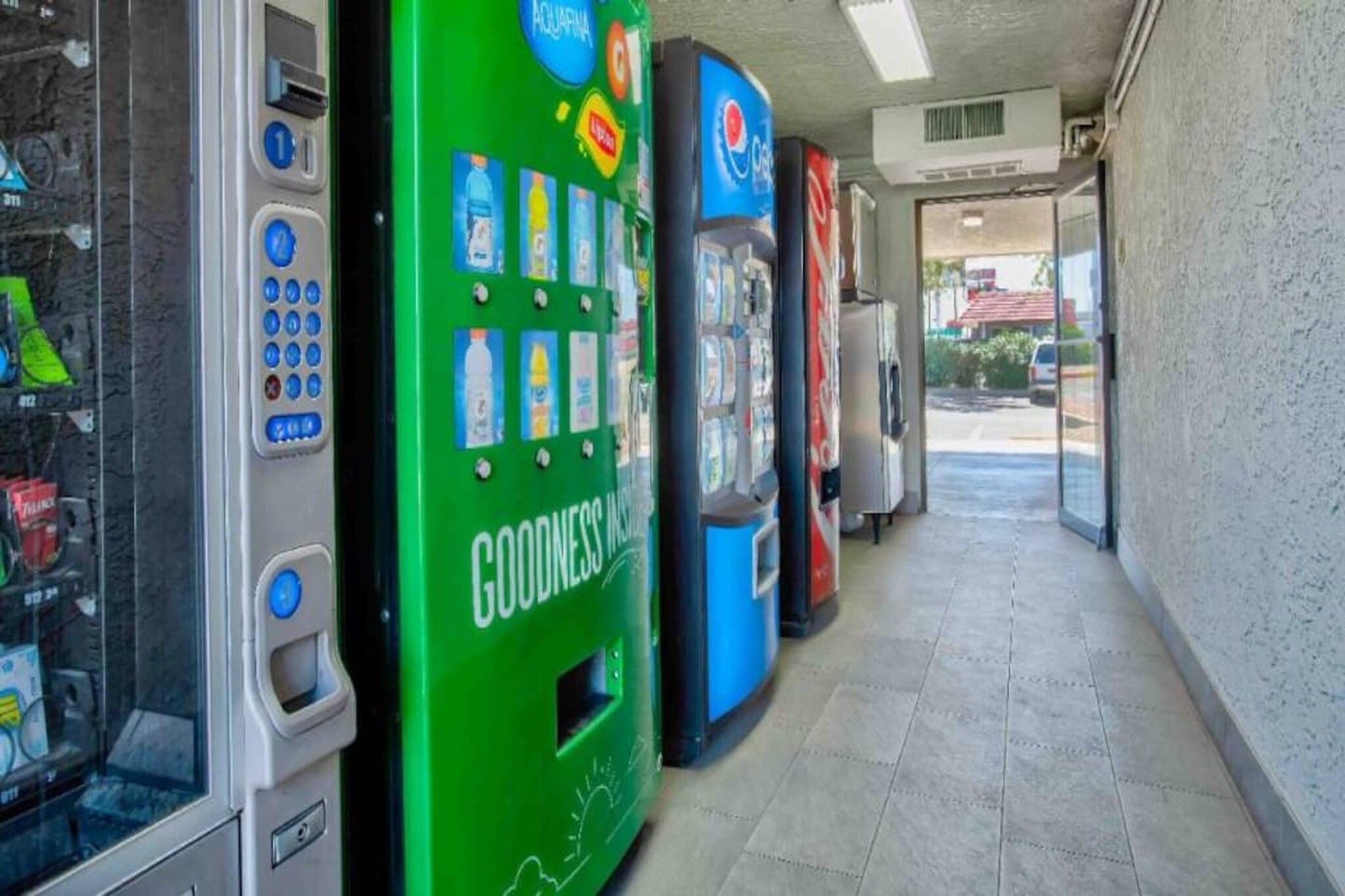 A row of vending machines in a commercial setting with a tiled floor and a view of the outside through a doorway.