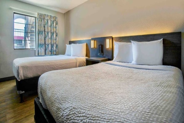 A hotel room with two beds, a window with patterned curtains, and a nightstand with a lamp. - 2 Queen Beds | Mini Fridge (Kingman)