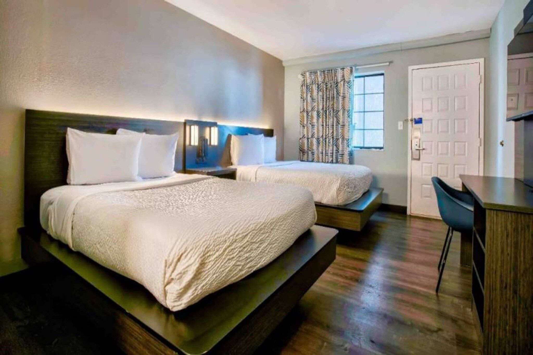 A hotel room with two beds, a desk, and a window with patterned curtains.