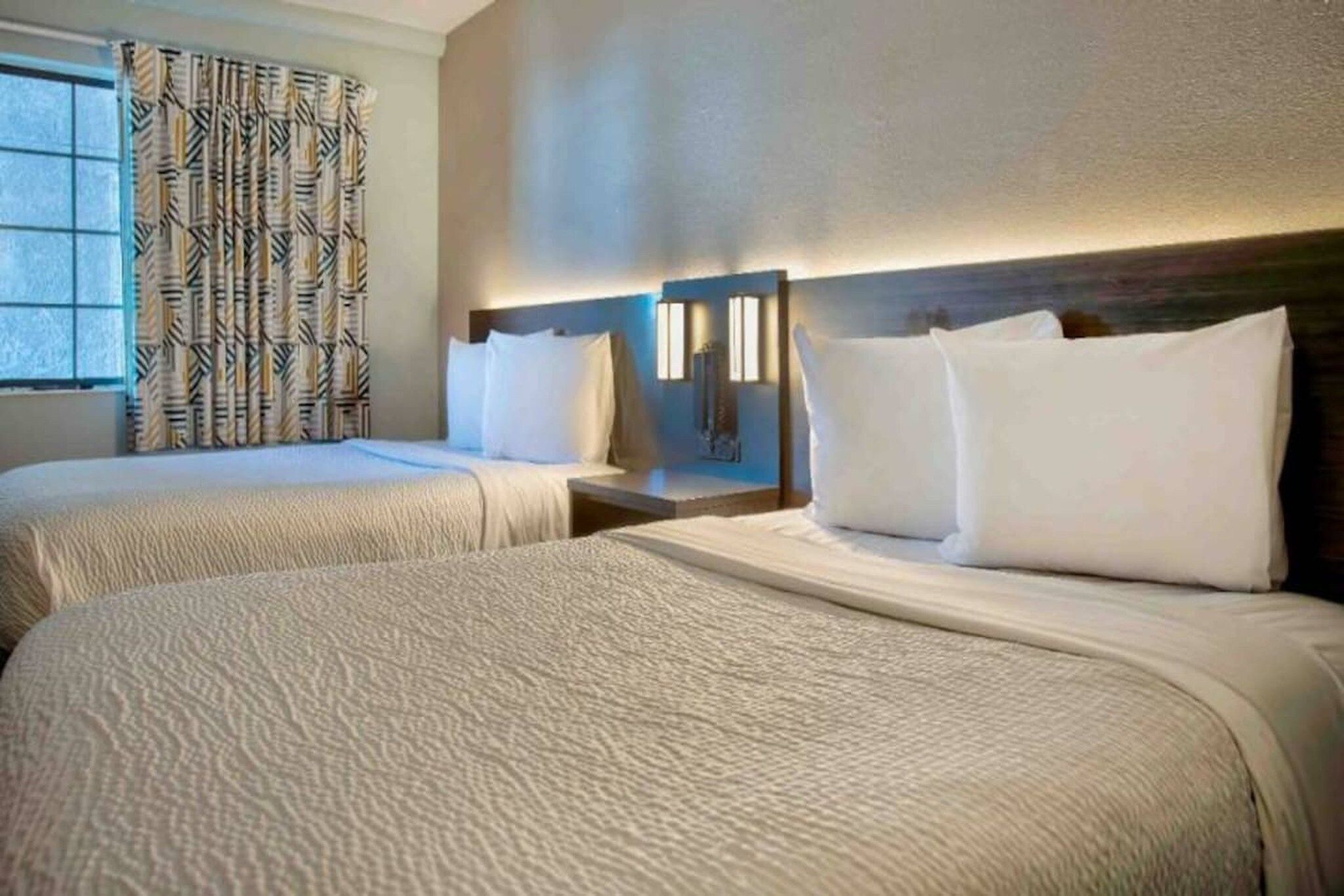 A hotel room with two beds, a window with patterned curtains, and a wall-mounted light fixture.