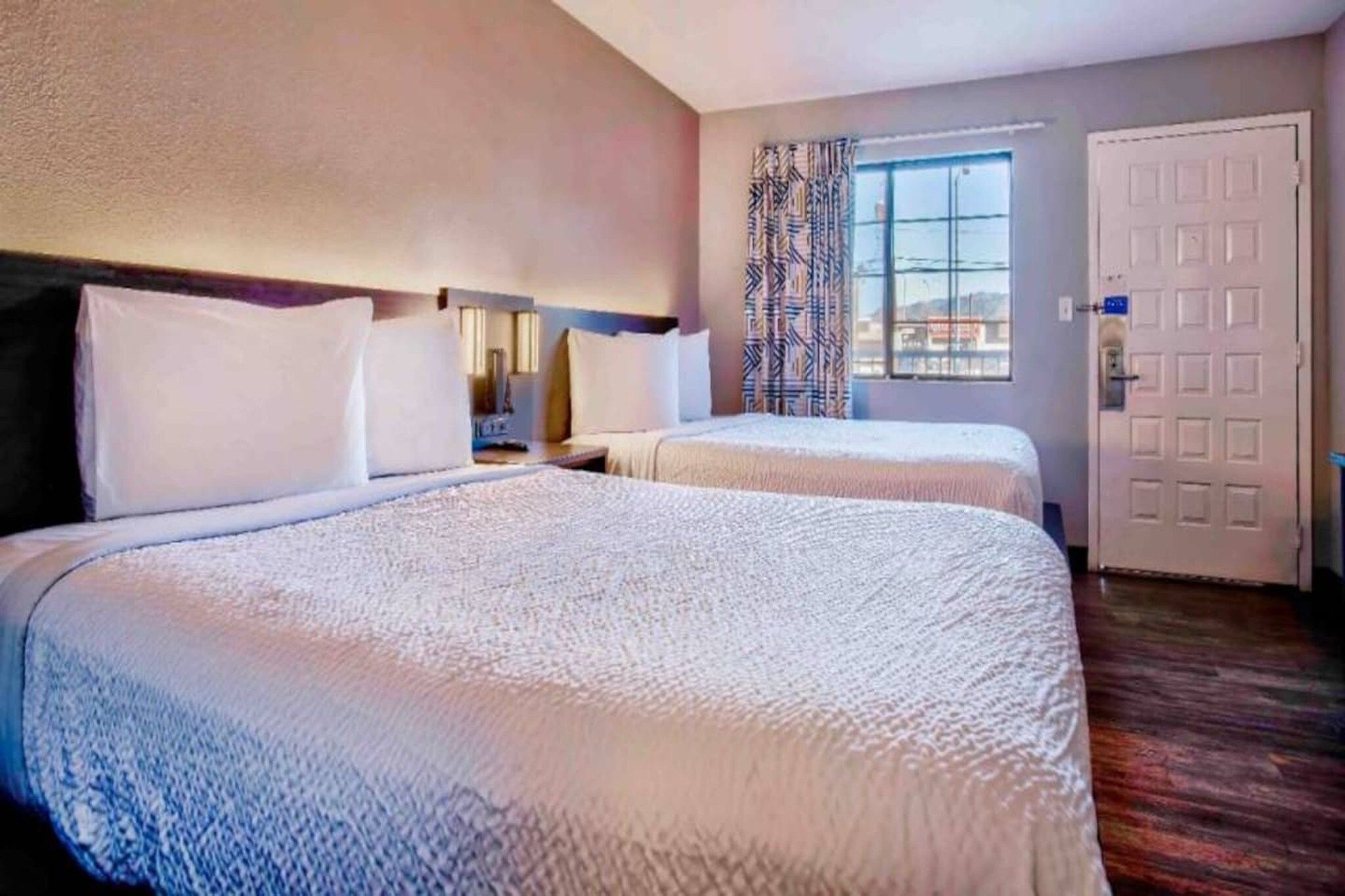 A hotel room with two beds, a window with curtains, a door, and a nightstand.