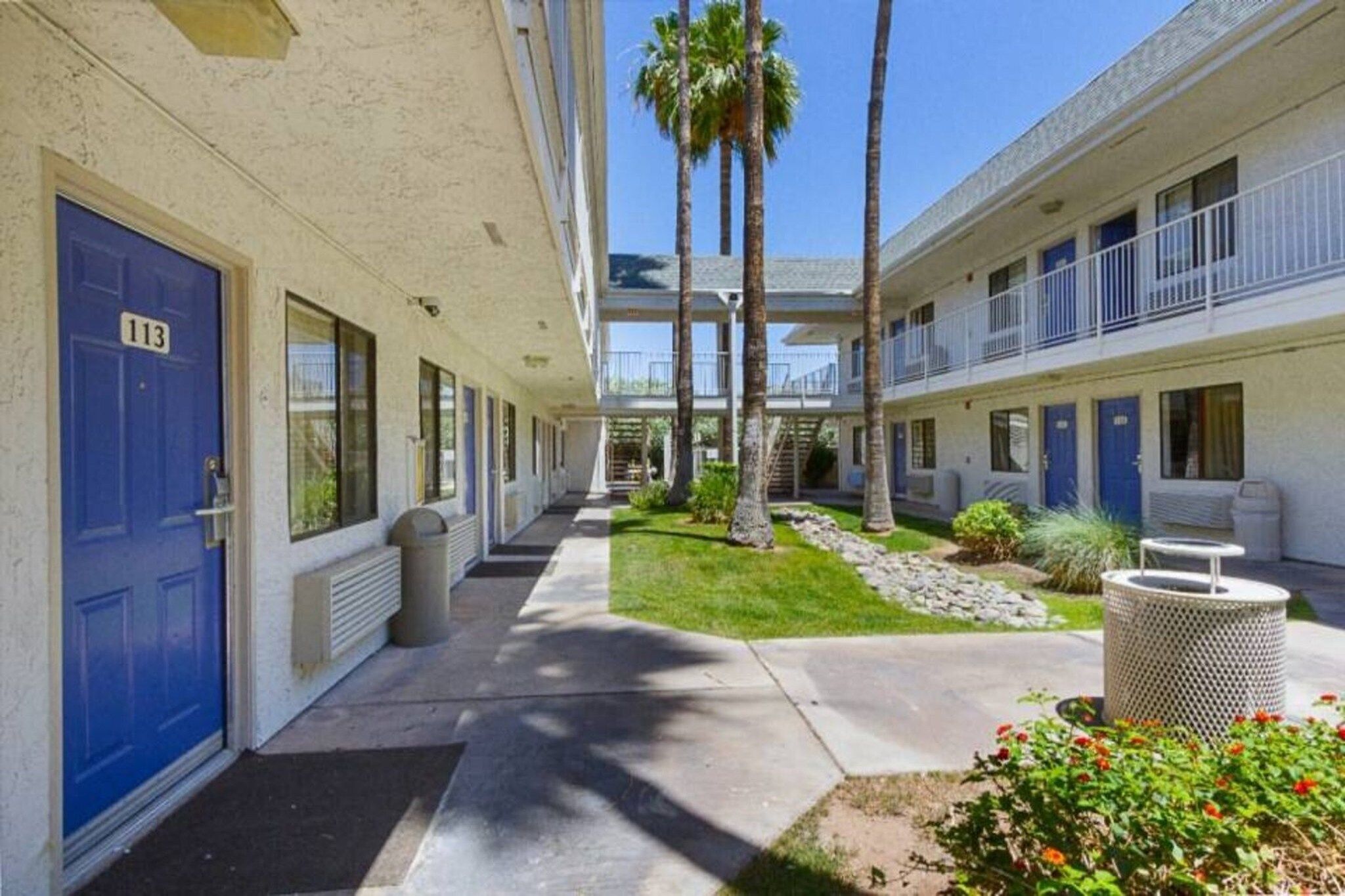 A hotel corridor with blue doors, a covered walkway, and a small garden area with palm trees and shrubs.