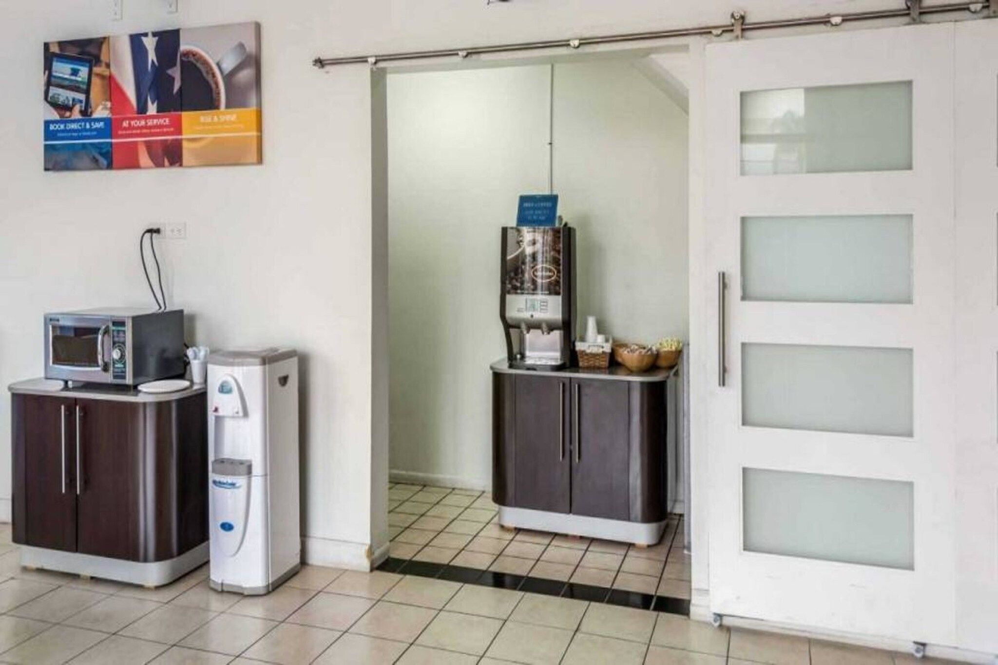 A kitchenette with a microwave, coffee maker, and a water dispenser. There is a cabinet with a coffee machine and a basket of bread. A door with frosted glass panels is visible.