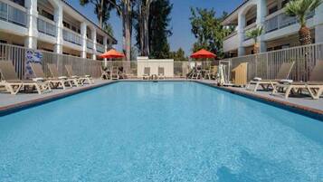 A swimming pool with lounge chairs and umbrellas, surrounded by a fence, and adjacent to a building with balconies.