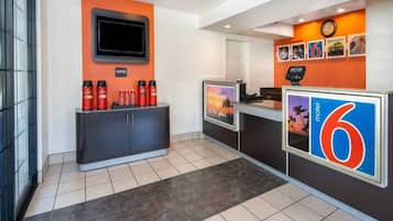 A hotel reception area with a coffee station, a TV screen, and a counter with a number sign.