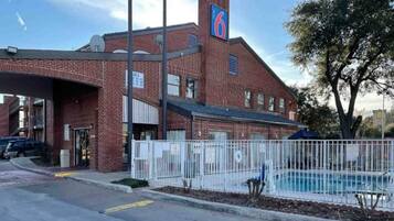 A brick building with a large sign on the roof, a fenced pool area, and a parking lot.
