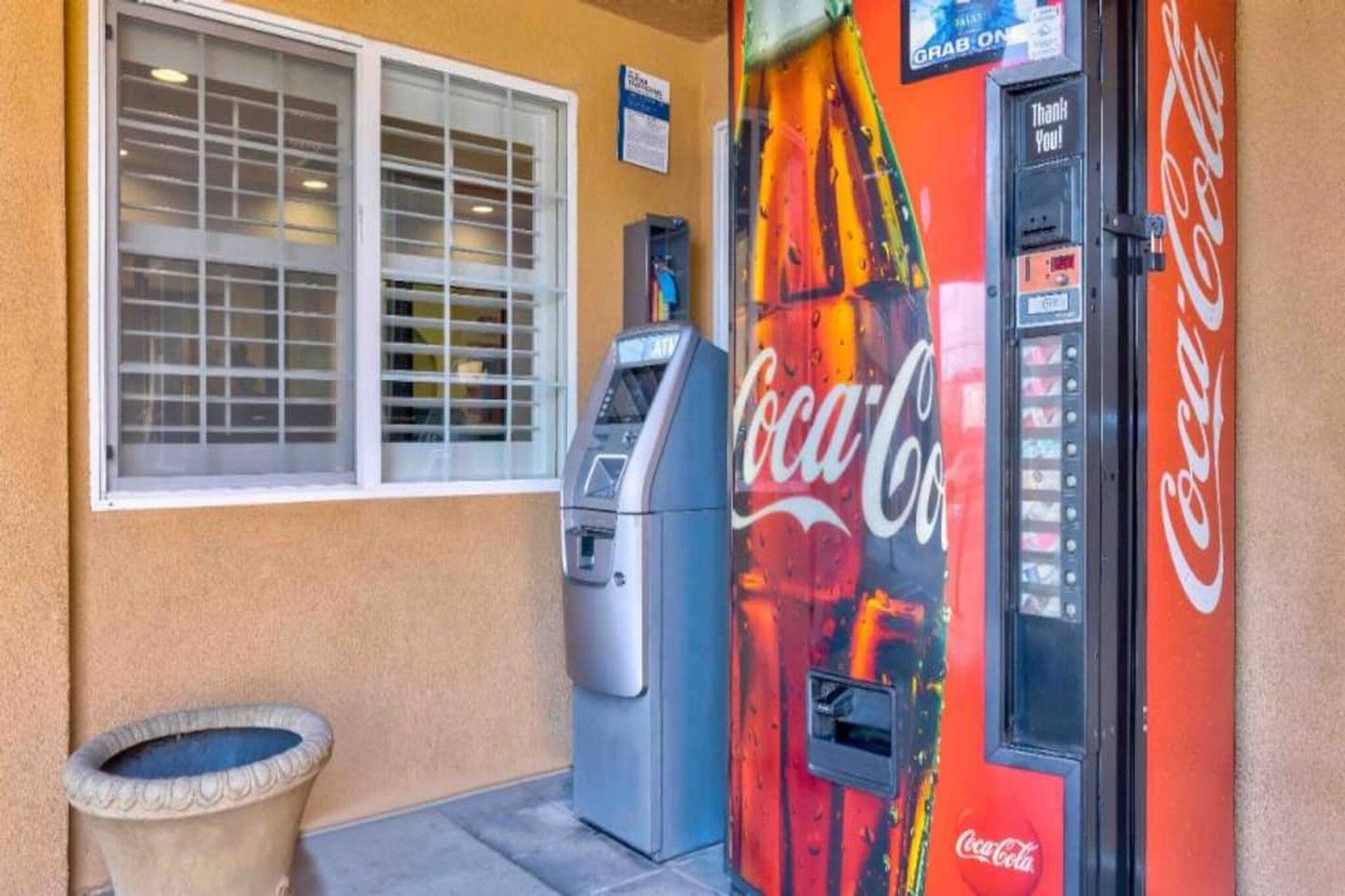 A Coca-Cola vending machine, a window with blinds, and a trash can outside a building.