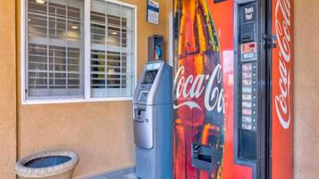 A Coca-Cola vending machine, a window with blinds, and a trash can outside a building.