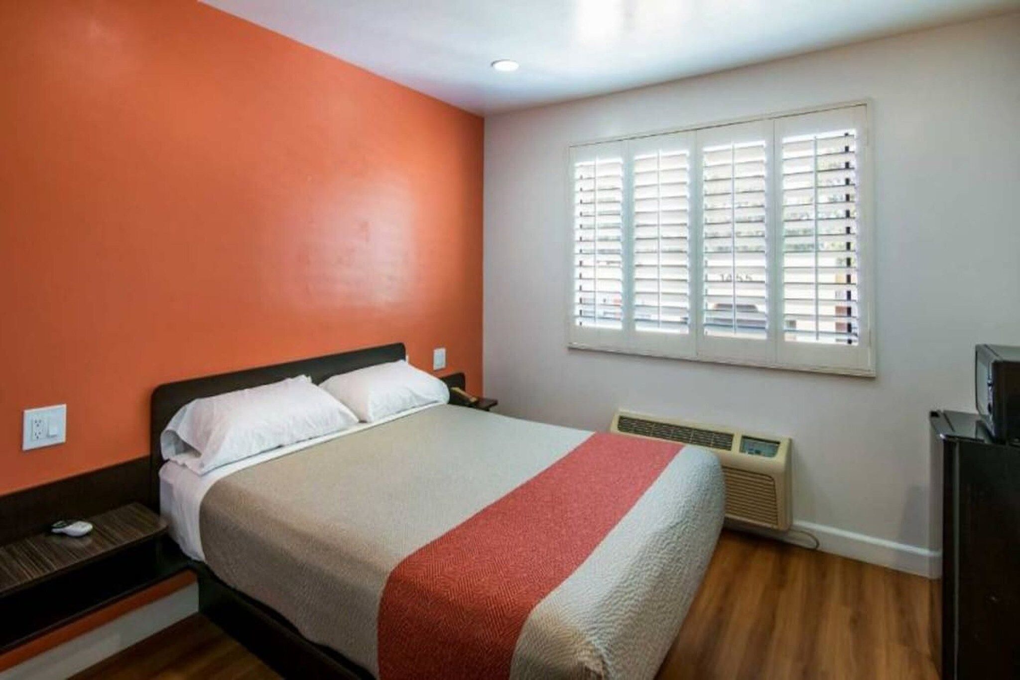 A hotel room with a bed, a heater, a window with blinds, and a wall with a red accent.