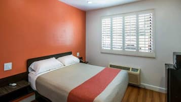 A hotel room with a bed, a heater, a window with blinds, and a wall with a red accent.