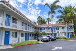 A motel with a two-story structure, multiple rooms with blue doors, and a parking area with cars. - Comfy 1 Queen Bed | Mini Fridge (Costa Mesa)