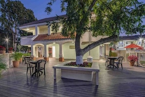 A hotel with a covered outdoor area featuring a dining table and chairs, surrounded by potted plants and a large tree.