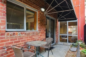 A brick building with a glass door entrance, a small outdoor seating area with wicker chairs and a table, and a potted plant on the right. - Barber`s Hyde | 2BR, Walk to Cafés & Waterfront (Lindisfarne)