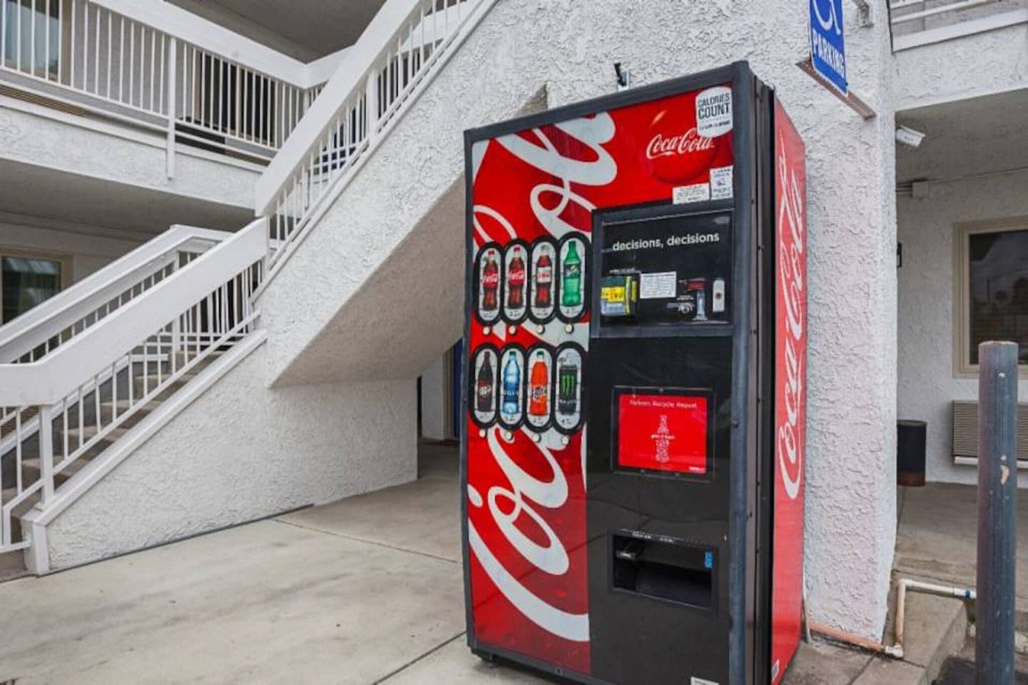 A Coca-Cola vending machine with a selection of beverages, located outside a building with a staircase.