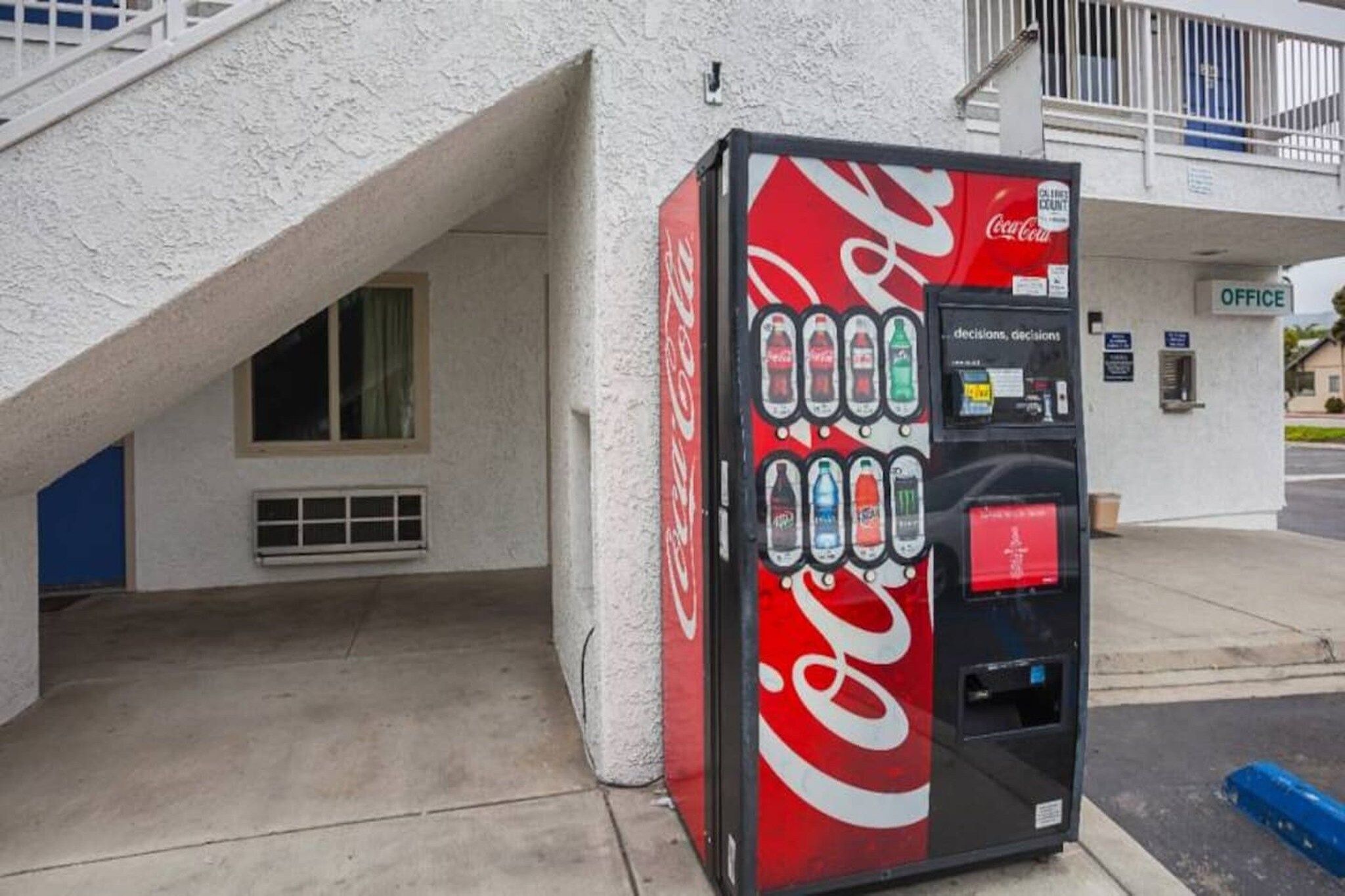 A Coca-Cola vending machine with a selection of beverages, located outside a building with an office sign.