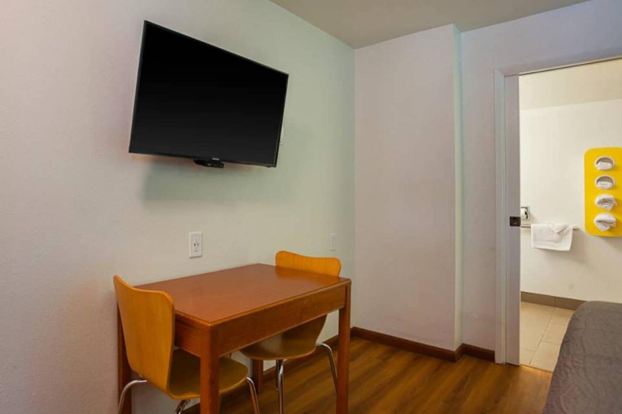 Une pièce dotée d’un téléviseur à écran plat fixé au mur, d’une table en bois avec deux chaises et d’une porte donnant sur une salle de bain équipée d’un porte-serviettes.